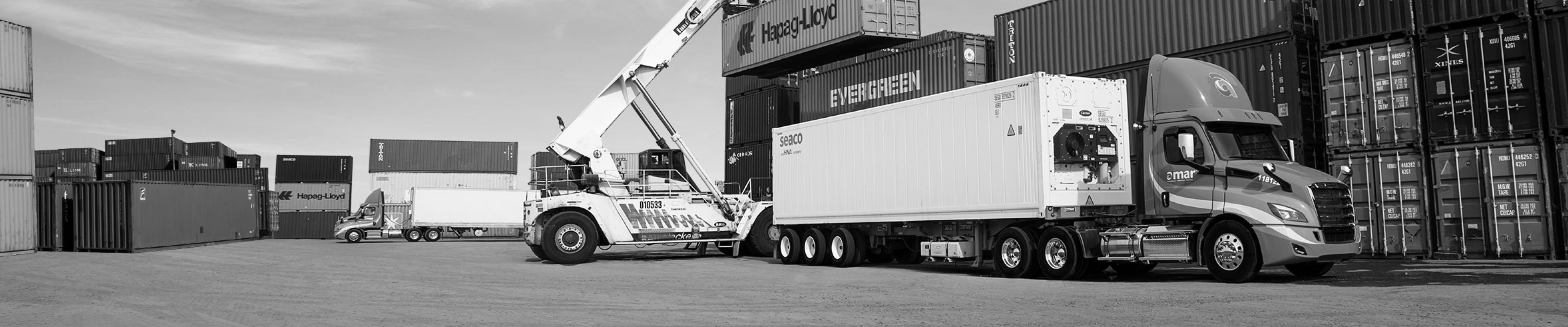 Black and white photo of Amar truck in container yard with reach stacker crane lifting a container behind it
